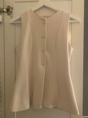 Beige väst med knappar från H&M - Säljer en stilren beige väst från H&M i storlek XS. Västen har en rak passform, rund halsringning och fyra klädda knappar framtill. Materialet känns mjukt och har en lätt struktur, perfekt för att lyfta vilken outfit som helst.