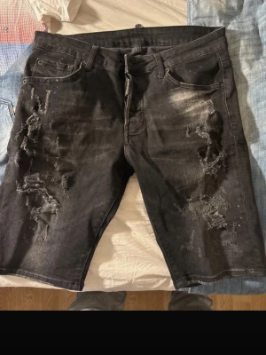 Svarta Dsquared2 shorts - Säljer ett par svarta jeansshorts med slitna detaljer och råa hål på båda benen. De har klassisk femficksdesign och dragkedja framtill. Perfekta för dig som gillar en edgy och avslappnad stil.