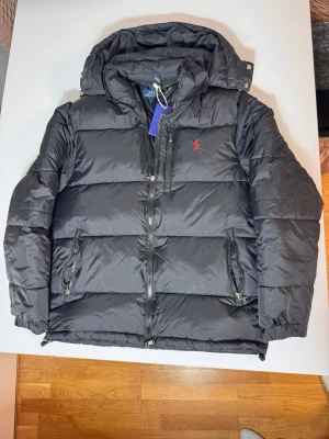 Svart pufferjacka från Polo Ralph Lauren - Säljer en svart pufferjacka från Polo Ralph Lauren med huva och dragkedja. Jackan har det klassiska röda Polo-logot broderat på bröstet och flera praktiska fickor med dragkedja. Perfekt för kalla dagar och riktigt snygg till streetwear-stilen.