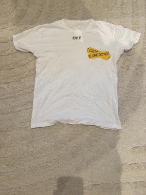 Vit Off-White t-shirt med tryck - Säljer en vit t-shirt från Off-White med korta ärmar. Framtill finns ett litet svart tryck och ett gult grafiskt tryck med texten 'FIRE LINE DO NOT CROSS'. Baksidan har ett stort gult tryck i form av korsande band med samma text. Materialet är mjuk bomull.
