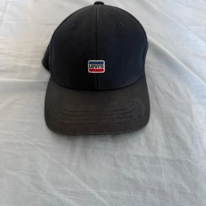 Svart keps från Levi's - Snygg svart keps från Levi's med klassisk böjd skärm och broderad logotyp framtill i rött, vitt och blått. Kepsen har justerbar rem baktill och är tillverkad i bomull för en bekväm passform. Perfekt accessoar för en avslappnad streetstyle.