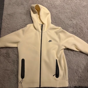Beige Nike hoodie med dragkedja - Snygg beige hoodie från Nike med svart dragkedja och svarta detaljer vid fickorna. Jackan har huva och Nike-logga på bröstet. Perfekt för chill eller streetwear, med lång ärm och stilren design.