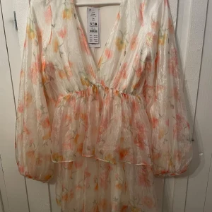 Blommig kortklänning med volang - Superfin kortklänning i ljus material med blommigt mönster i rosa, orange och gult. Klänningen har v-ringning, långa puffärmar och volangdetaljer i kjolen. Perfekt för sommarens alla tillfällen! Helt ny med prislapp kvar! Pris kan diskuteras.