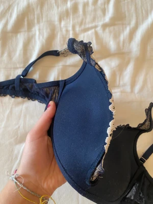 Intimissimi bh - Säljer en mörkblå bh med vacker spets och broderade blommor i vit. Axelbanden är justerbara och har spetsdetaljer, och kanten är dekorerad med broderad spets. Kuporna är vadderade och ger snygg form. Push up från intimissimi. Nästan inte använd köpt för någon vecka sedan, storlek 75b 