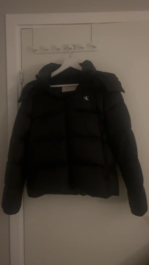Svart pufferjacka Calvin Klein Jeans - Svart pufferjacka från Calvin Klein Jeans med klassisk CK-logga på bröstet. Jackan har hög krage, långa ärmar och dragkedja framtill. Själv brukar jag ha S i tröjor och denna jacka passar perfekt för mig. 