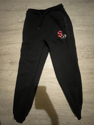 Svarta mjukisbyxor med röd logga - Svarta mjukisbyxor med snörning i midjan och en röd och vit SD-logga på vänster lår. Byxorna har ribbade muddar vid bensluten och är gjorda i mjukt material som passar chill och streetwear. Perfekt för att hänga med kompisar eller bara ta det lugnt.