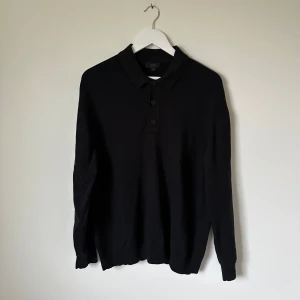 Svart pikétröja från COS i bomull - Svart långärmad pikétröja från COS i 100% bomull. Klassisk krage och tre knappar framtill. Tröjan har en stilren och enkel design som passar perfekt till jeans eller chinos. Mjuk och skön kvalitet med normal passform. 5J6