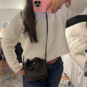 Svart croco axelväska med gulddetalj - Snygg svart axelväska i croco-mönstrat skinnimitation med guldfärgad knapp framtill. Väskan har både handtag och lång axelrem, perfekt storlek för det viktigaste. Stilren och trendig design som lyfter din outfit.