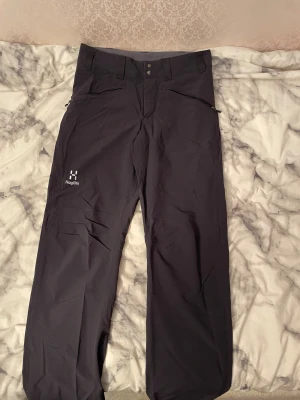 Svarta täckbyxor från Haglöfs herr S - Svarta täckbyxor från Haglöfs i herrmodell, storlek S. Byxorna har två dragkedjeförsedda fickor framtill, bälteshällor och tryckknappar i midjan. Materialet är slitstarkt syntet, perfekt för utomhusaktiviteter under kyliga dagar.