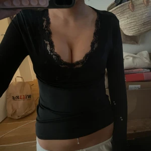 Svart långärmad topp med spets - Säljer en svart långärmad topp med djup v-ringning och snygga spetsdetaljer runt halsen. Toppen är tight och har en feminin look som passar perfekt till jeans eller kjol. Superenkel att styla för en trendig outfit.