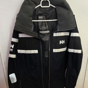 Svart Helly Hansen vindjacka/seglarjacka S - Svart vindjacka från Helly Hansen med vita reflexdetaljer och logga på bröstet och ärmen. Jackan har hög krage, kapuschong,dragkedja och meshfoder. Perfekt för blåsiga dagar och har en sportig look med tydliga kontraster. Strl.S/P. Helly Tech protection