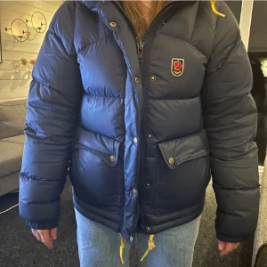 Blå dunjacka från Fjällräven - Säljer en blå dunjacka från Fjällräven med gula snören💙 Jackan har två stora fickor med lock, huva och klassisk Fjällräven-logga på bröstet. 
