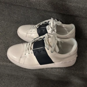 Valentino Garavani Open sneakers vit/blå - Snygga Valentino Garavani Open sneakers i vitt skinn med bred mörkblå rand över mitten. Klassisk rund tå, platt sula och vita snören. Ikoniska nitar baktill ger extra edge. Perfekta för dig som gillar stilrena och lyxiga sneakers. Pris kan diskuteras! Nya skor