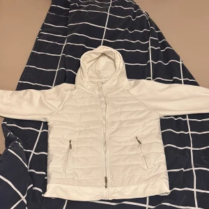 Vit cardigan pufferjacka med luva cilver dragkedjor - Snygg vit pufferjacka från JLAY med huva och dragkedja framtill. Jackan har quiltad framsida, två fickor med dragkedja och släta ärmar. Perfekt för dig som gillar stilren och clean look. Passar dig som vill ha en jacka med normal passform och lång ärm. Stil grisch jacka