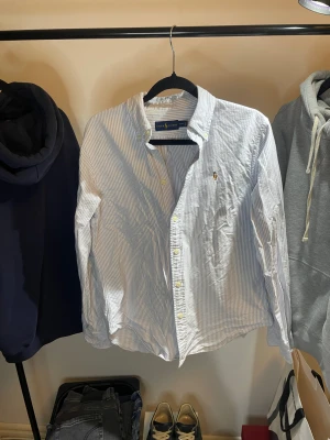 Randig skjorta från Ralph Lauren  - Snygg skjorta från Ralph Lauren i klassisk vit och ljusblå randig design. Skjortan har lång ärm, knappar framtill och ikoniska broderade loggan på bröstet. Tillverkad i mjuk bomull som känns skön mot huden. Perfekt för dig som gillar stilrena och fräscha plagg.