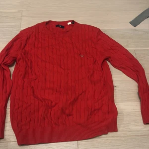 Röd kabelstickad tröja från GANT - Snygg röd kabelstickad tröja från GANT med rund hals och ribbade muddar. Tröjan har ett litet broderat GANT-logo på bröstet och är långärmad. Perfekt för dig som gillar klassisk stil med en modern touch.