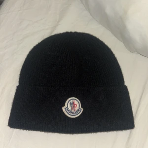 Svart Moncler mössa i ull - Svart ribbstickad mössa från Moncler med klassisk rundad form och uppvikt kant. Framtill sitter Monclers ikoniska broderade logotyp i vitt, blått och rött. Mössan är tillverkad i mjuk ull och passar perfekt för kyliga dagar.