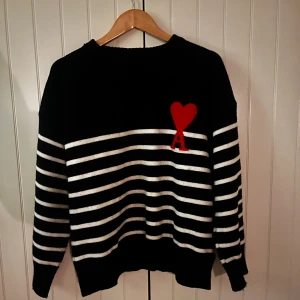Mörkblå och vit randig tröja från Ami - Mörkblå stickad tröja från Ami med vita horisontella ränder och en röd hjärtapplikation med bokstaven A på bröstet. Tröjan har långärmad passform och är tillverkad i mjukt stickat material. Perfekt för dig som gillar stilrena detaljer med en twist. Tröjan ser svart ut men den är egentligen en väldigt mörk mörkblå 