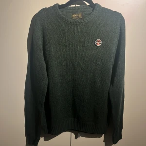 Mörkgrön stickad tröja Morris - Jag säljer en väldigt fin mörkgrön stickad tröja från Morris med rund hals och ribbade muddar. Tröjan har ett snyggt Morris-emblem på bröstet och är tillverkad i mjuk bomull. Perfekt för dig som gillar klassisk stil med en modern touch.