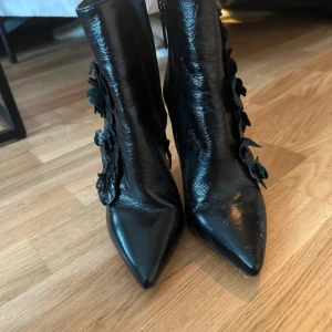 Svarta stövletter med blommor och klack från Zara - Säljer ett par svarta stövletter från Zara i äkta läder med hög klack och spetsig tå. Skorna är gjorda i glansigt skinn och har dekorativa blommor på utsidan som ger en unik och cool vibe. Dragkedja på insidan för enkel påtagning. Perfekt för dig som vill sticka ut.