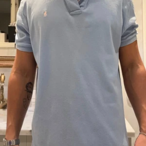 Ljusblå polo från Ralph Lauren - Snygg ljusblå pikétröja från Polo Ralph Lauren i storlek M. Klassisk krage, korta ärmar och knappar vid halsen. Ikoniska broderade loggan på bröstet. Mjuk bomull som känns skön mot huden och passar perfekt till jeans eller shorts.