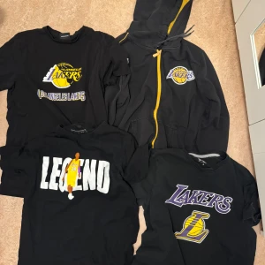 Svart Lakers hoodie med gul dragkedja - Svart hoodie från Los Angeles Lakers med gul dragkedja och huva. Lakers-logga broderad på bröstet och gula detaljer längs sömmarna. Perfekt för dig som älskar basket och vill ha en sportig streetstyle. Mjuk och bekväm med fickor framtill.