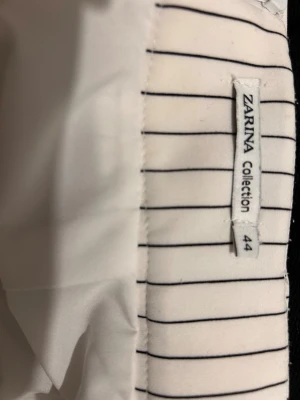 Vit randig långkjol från Zarina - Snygg vit långkjol från Zarina Collection med svarta vertikala ränder och ett matchande bälte i midjan. Kjolen har en slits framtill som ger extra edge och är tillverkad i ett mjukt polyestermaterial. Perfekt för dig som gillar stilrena och moderna plagg. EU-storlek 38 (står det 44)