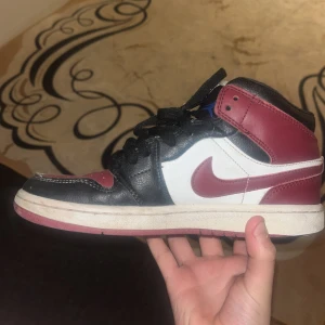 Nike Air Jordan 1 Mid  (obs äkta) - Nike Air Jordan 1 Mid i svart, rött och vitt skinn. Klassisk siluett med röd sula, svart snörning och blå Jumpman-logga på plösen. Snygg kontrast mellan färgerna och ikonisk design som alltid är rätt.