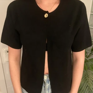 Svart bolerotopp med guldknapp - Säljer en svart bolerotopp med korta ärmar och rund halsringning. Toppen har en snygg guldknapp framtill som stängning och är öppen nedtill. 