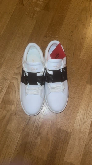 Valentino Garavani Open sneakers vit/svart - Snygga Valentino Garavani Open sneakers i vitt skinn med bred svart läderdetalj över vristen. Klassisk rund tå, platt sula och vita snören. Ikonisk och stilren design som lyfter vilken outfit som helst.