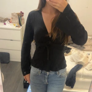 Svart omlottopp med knytband - Säljer en svart omlottopp med långa ärmar och v-ringning. Toppen har ett knytband i midjan som ger en snygg siluett och är tillverkad i ett mjukt tyg. Perfekt att styla med jeans eller kjol.