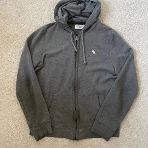 Grå hoodie Abercrombie & Fitch M - Grå hoodie från Abercrombie & Fitch i storlek M. Tröjan har huva med snörning, dragkedja framtill och två fickor. Liten vit logga med älg broderad på bröstet. Mjuk bomullsmix som är skön att ha på sig och passar perfekt till chill dagar.