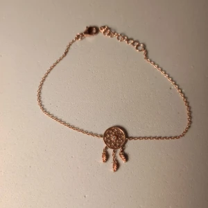 Roséguldfärgat armband med drömfångare - Snyggt armband i roséguld med en detaljrik drömfångare i mitten och tre små hängande fjädrar. Kedjan är tunn och justerbar för perfekt passform. Perfekt accessoar för dig som gillar boho-stil och unika smycken. 