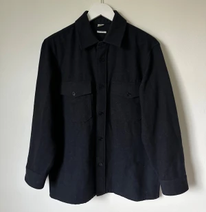 Grå overshirt från Arket i ullmix - Grå overshirt från Arket i ullblandning med twillstruktur. Regular fit med klassisk krage, två bröstfickor med knappar och långa ärmar. Perfekt lager-på-lager-plagg med stilren look och snygga detaljer. 4A5