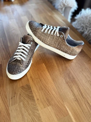 Bruna flätade sneakers från Hamlet - Snygga sneakers från Hamlet i brun nyans med flätad skinnstruktur och vita snören. Skorna har rund tå och vit platt sula som ger en clean look. Perfekt för dig som gillar detaljer och vill sticka ut med ett unikt mönster. Nyrpis 4500kr