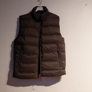 Grå dunväst från H&M, storlek M - Helt oanvänd grå dunväst från H&M med hög krage och två praktiska fickor med dragkedja. Västen har en quiltad design och stängs med dragkedja framtill. Perfekt för lager-på-lager och enkel att matcha med hoodies eller tröjor. Prislappen sitter även kvar.