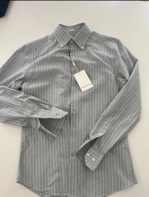 Randig slim fit skjorta These Glory Days Oxford skjorta  - Snygg randig skjorta från These Glory Days i slim fit-modell. Skjortan är ljusgrå med vita och svarta ränder, har klassisk krage och vita knappar framtill samt vid ärmslut. Tillverkad i mjuk bomull, perfekt för dig som gillar stilrena och fräscha plagg. 