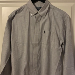 Randig skjorta från Polo Ralph Lauren - Snygg randig skjorta från Polo Ralph Lauren i klassisk design med button-down krage och broderad logga på bröstet. Skjortan är vit med tunna grå ränder, har lång ärm och knäppning framtill. Perfekt för dig som gillar stilrena och tidlösa plagg.