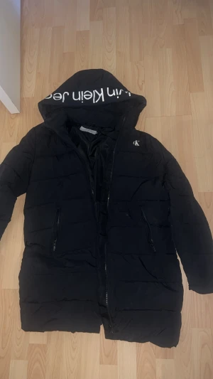 Svart pufferjacka från Calvin Klein Jeans - Svart pufferjacka från Calvin Klein Jeans med stor huva och vit logga på insidan. Jackan har två sidofickor med dragkedja, quiltad design och CK-brodyr på bröstet. Perfekt för kalla dagar med sin långa modell och snygga streetstil.