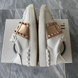 Valentino Rockstud sneakers vit/guld - Snygga Valentino Rockstud sneakers i vitt skinn med guldiga detaljer och ikoniska nitar. Skorna har platt sula, rund tå och snörning. Perfekta för dig som vill sticka ut med exklusiva detaljer och cool design.