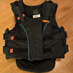  Rid Svart säkerhetsväst från Airowear - Säljer en svart säkerhetsväst från Airowear med blå detaljer och justerbara kardborreband. Västen har perforerat material för ventilation och är designad för ridning. Märket syns tydligt både fram och bak. Perfekt för dig som vill ha extra skydd vid hästaktiviteter.