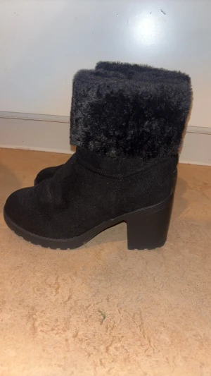 Svarta boots med fuskpäls och klack - Snygga svarta boots med hög klack och grov sula. Skorna har mjuk fuskpäls upptill och är tillverkade i mockaliknande material. Dragkedja på sidan gör dem enkla att ta på. Perfekta för kalla dagar när du vill vara både varm och stilren.