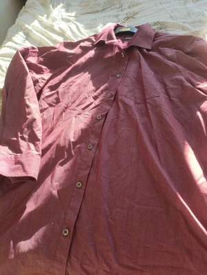 Vinröd skjorta från Dressmann - Snygg vinröd skjorta från Dressmann med klassisk krage och knappar hela vägen. Skjortan har lång ärm och ett diskret bröstficka. Materialet känns som bomull och har en lätt melerad look. Perfekt för dig som gillar stilrena plagg med lite färg.