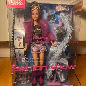 Barbie Fashion Show klänning med paljetter från 2004 - Säljer en Barbie Fashion Show klänning från Mattel med coola detaljer! Klänningen är kort, svart och rosa med glittriga paljetter och stjärnmönster, samt en lila fluffig jacka. Ingår även en lång silverfärgad klänning med paljetter och glittriga detaljer. Perfekt för dig som älskar glam och mode! Från 2004