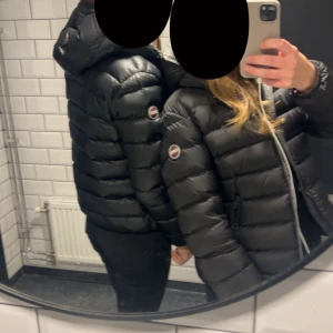 Colmar jacka  - Säljer en svart colmar jacka  med huva och snygga breda kanaler. Jackan har en rund logotyp på ärmen och dragkedja framtill. Perfekt för dig som vill ha en stilren och varm jacka med modern look.
