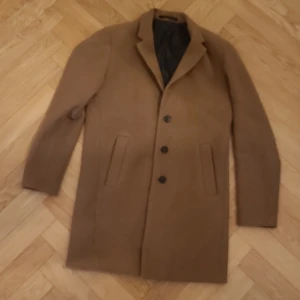 Rock  från Jack & Jones M - Snygg beige rock från Jack & Jones Premium i storlek M. Klassisk design med tre knappar framtill, två fickor och krage. Tillverkad i ullblandning med mörkt foder. Perfekt för dig som vill ha en stilren och tidlös look.