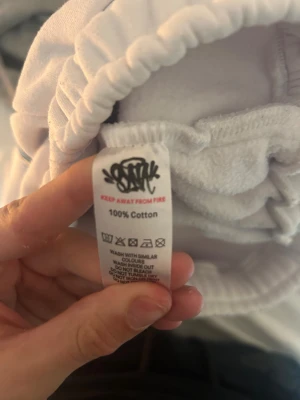 Vita sweatpants från syna world  - Säljer ett par vita sweatpants från syna world  med elastisk midja och stort blått graffiti-tryck på baksidan. På ena benet finns broderad text 'Forgive Me' i ljusblått. Tillverkade i 100% bomull för skön känsla. Perfekt för chill dagar och streetwear-vibe.