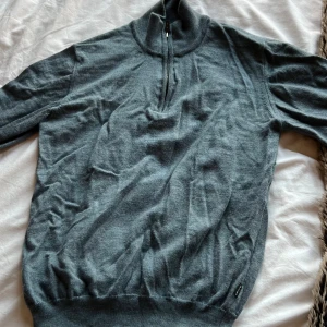 Grå half zip stickad tröja Sand - Snygg grå stickad tröja från Sand Copenhagen med half zip och hög krage. Insidan av kragen har ett blommigt tyg som ger en unik detalj. Perfekt för dig som gillar stilrena plagg med en twist. Tröjan har långa ärmar och ribbade muddar.