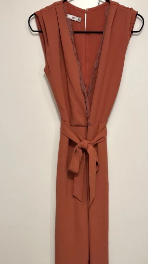 Byxdress från MANGO - Säljer en snygg mörkrosa byxdress från MNG med knytband i midjan och spetsdetaljer längs den djupa v-ringningen. Byxdressen har vida ben och är ärmlös.