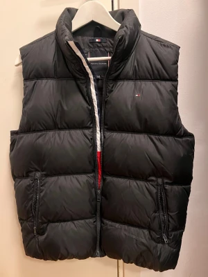 Svart dunväst från Tommy Hilfiger - Snygg svart dunväst från Tommy Hilfiger med hög krage och dragkedja framtill. Västen har två fickor med dragkedja och ett stort Tommy Hilfiger-tryck i vitt och blått på ryggen. Perfekt för lager på lager och streetwear-vibe.
